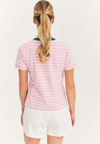 Kobieta z blond warkoczem, ubrana w różowo-białą koszulę w paski z zielonym kołnierzykiem i białe szorty, zwrócona plecami na jednolitym tle.