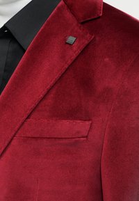 Μπορντό βελούδινο σακάκι με πέτο με εγκοπή, τσέπη στο στήθος και μαύρο γιακά πουκάμισου από κάτω, με μια μικρή μαύρη τετράγωνη καρφίτσα στην πέτο.