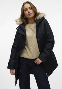 Melnā polsterētā parka ar mākslīgās kažokādas kapuci, ar rāvējslēdzēja kabatām un priekšējo aizdari, uzvilkta pār gaiši bežīgu teksturētu adītu džemperi.