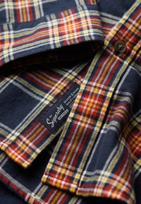 Camisa de franela a cuadros con patrones en azul marino, rojo, amarillo y blanco. Lleva una etiqueta con "Superdry" y detalle de botones.