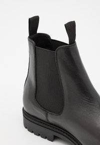 Botín Chelsea de cuero negro con panel elástico lateral, lengüetas en la parte delantera y trasera, y suela robusta con tacos sobre un fondo blanco.