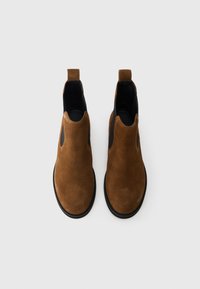 Καφέ σουέτ μποτάκια μέχρι τον αστράγαλο με μαύρες ελαστικές πλαϊνές πάνελ και θηλιά στο τακούνι. Μαύρη επίπεδη σόλα και στρογγυλή μύτη.