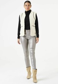 Crème kleurige fluffy vest met ritsdetails, gecombineerd met glanzende zilveren skinny jeans en beige enkellaarsjes. Zwarte coltrui eronder.