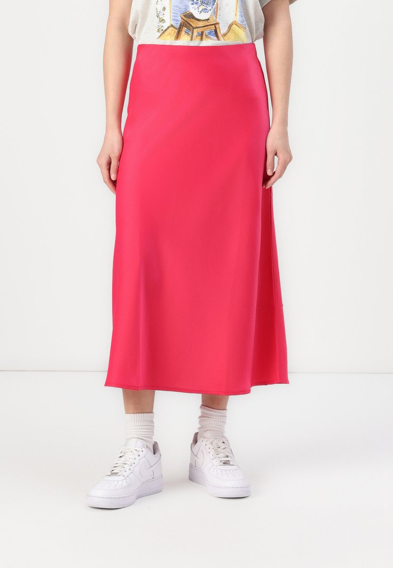 Jupe midi rose en tissu lisse, arborant un design simple et fluide. Associée à des baskets blanches et légèrement froncée à la taille.