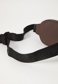 Καφέ υφασμάτινη τσάντα με μαύρη ρυθμιζόμενη λωρίδα που διαθέτει ανάγλυφο λογότυπο. Λείο υφή με καμπύλο, συμπαγές σχέδιο.