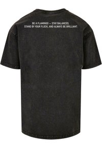 Camiseta de algodón negra de manga corta. La parte posterior cuenta con texto blanco: "SÉ UN FLAMINGO – MANTENTE EN EQUILIBRIO, PERMANECE JUNTO A TU BANDADA Y SIEMPRE SÉ BRILLANTE."