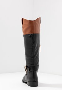 Bota alta de cuero negro y marrón con cremallera lateral y hebilla decorativa sobre un fondo blanco, mostrada desde la parte trasera y en ángulo lateral.