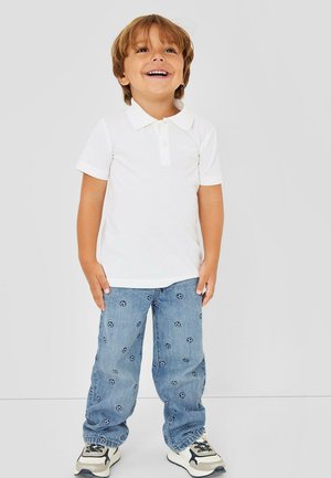 Glimlachend kind staand, draagt een wit polohemd, blauwe spijkerbroek met kleine voetbalprints en witte sneakers met zwarte details.