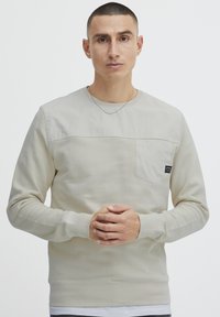 Beige színű, hosszú ujjú pulcsi, kerek nyakkal és mellzsebel. Jellemzője a textúrázott felső panel és bordás mandzsetta. Minimalista dizájn.