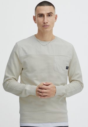 Sudadera beige de manga larga, cuello redondo y bolsillo en el pecho. Presenta un panel superior texturizado y puños acanalados. Diseño minimalista.