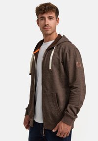 Brown zip-up hoodie with a textured finish, cream drawstrings, side pockets, and a logo patch on the sleeve. Layered over a white shirt.