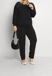 Svart sweatshirt med nitar på axlarna, parat med svarta byxor och vita ballerina skor. En svart handväska hålls av modellen. Enkelt design.