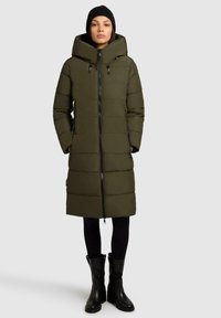 Kvinna som bär en lång jacka i olivgrön vadderad vinterjacka med huva, svart mössa, svarta leggings och svarta stövlar till mitten av vaden, står rak.