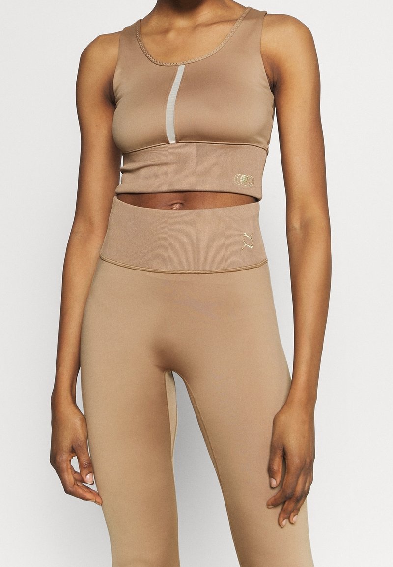 Femme portant un crop top beige et des leggings taille haute avec des détails de couture et des accents réfléchissants subtils, debout sur un fond uni.