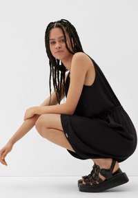 Schwarzes ärmelloses Kleid mit elastischem Taillenzug, strukturiertem Stoff und einem Logo-Tag. Schwarze Plattform-Sandalen mit überkreuzten Riemen.