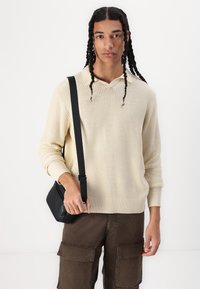 Bež pleteni pulover s V-izrezom, rebrastom teksturom i dugim rukavima; uparen s tamnosmešnim cargo hlačama i malom crnom torbom za crossbody.