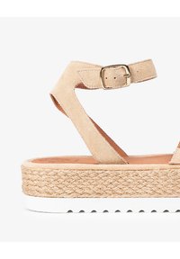 Sandalia de ante beige con tiras cruzadas, hebilla dorada y plataforma de yute trenzado. La suela de goma blanca presenta un diseño acanalado para mayor agarre.