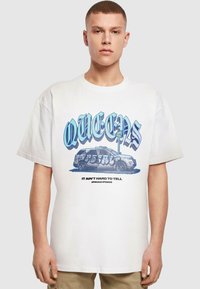 Fehér pamut póló, merész kék "QUEENS" grafikával, autóillusztrációval és az "IT AIN'T HARD TO TELL" felirattal, laza szabással.