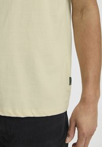 Camiseta de algodón de color beige claro con cuello redondo y dobladillo recto, con una pequeña etiqueta de marca negra en el costado, combinada con pantalones oscuros.