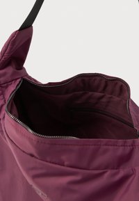 Bolsa de tela de nylon morada con cierre superior y bolsillo interior. Tiene una textura suave y una correa negra resistente.