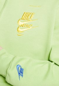 Ljusgr ön sweatshirt med gula och blå Nike-logotyper. Tyget ser mjukt ut med en slät textur och en åtsittande ärmdesign.