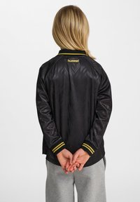 Svart sportjacka med mönstrad textur, gula detaljer på kragen och ärmarna, samt "hummel"-logotypen broderad på baksidan.