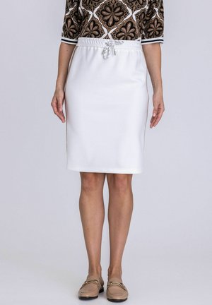 Frau trägt einen weißen knielangen Rock mit Kordelzug in der Taille, ein gemustertes schwarzes und braunes Oberteil sowie beige Slipper, steht vor einem schlichten Hintergrund.