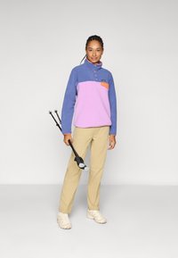 Fleece pullover in blue and pink with a high collar, orange pocket, and buttons. Light tan pants and beige shoes. Holding trekking poles.