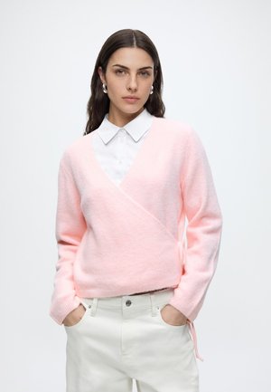 Femme aux longs cheveux foncés portant un pull enveloppant duveteux rose clair sur une chemise blanche à col, un pantalon blanc et des boucles d'oreilles créoles argentées.