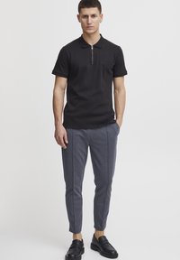 Polo-shirt noir avec un col zippé, associé à un pantalon gris ajusté avec des coutures latérales. Le modèle porte des mocassins noirs.