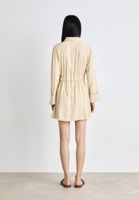 Robe chemise beige courte avec de longues manches, taille cintrée et jupe évasée. Tissu lisse, détails minimalistes, assortie à des sandales plates.