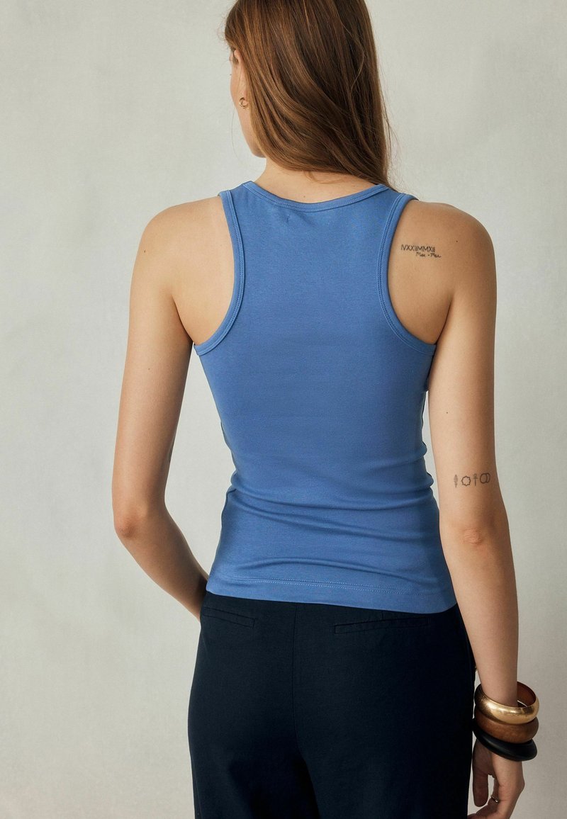 Mujer con el cabello largo y castaño, vestida con un top azul sin mangas y pantalones negros, mostrando tatuajes en el brazo izquierdo superior y el hombro derecho, de pie frente a un fondo liso.