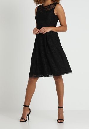 Kobieta ubrana w czarną koronkową sukienkę bez rękawów do kolan i czarne wysokie sandały na kostkę, stoi ze złączonymi rękami z przodu.