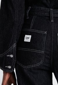 Bolsillo trasero y puño de una chaqueta de mezclilla negra y unos vaqueros con etiquetas de la marca Lee Workwear en blanco y botones plateados en el puño.