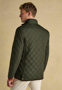 Mladý muž v tmavozelenej prešívanej bunde, stojaci bokom s väčšinou viditeľným chrbtom na jednoduchom béžovom pozadí.