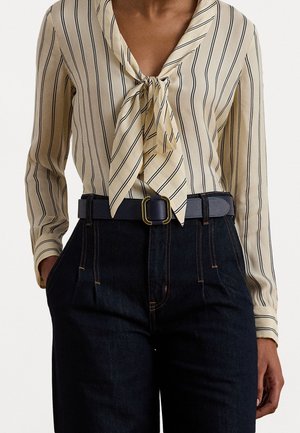 Personne portant un chemisier beige à manches longues avec des rayures noires et un nœud à l'avant, associé à un jean foncé taille haute et une ceinture noire avec une boucle dorée.