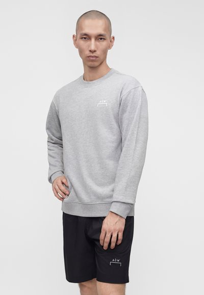 Άνδρας που φορά ανοιχτογκρι μακρυμάνικη μπλούζα και μαύρα σορτς με μικρά λευκά λογότυπα A-C-W, στέκεται μπροστά από ένα απλό ανοιχτό φόντο.