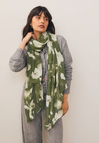 Sciarpa verde con motivo floreale, realizzata in tessuto leggero e strutturato, avvolta attorno al collo. Il tessuto presenta un disegno ondulato.