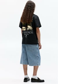 Sort grafisk t-shirt med "La Gioconda" kunstværk på ryggen, parret med lyseblå denimshorts og sorte sko med hvide sokker.
