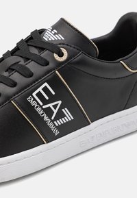 Svart syntetisk sneaker med vit sula. Har broderad "EA" logotyp, guldaccenter och snörningsdesign. Slät textur med eleganta linjer.