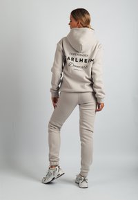 Conjunto de sudadera con capucha y pantalones de chándal beige, confeccionado con tejido suave, que presenta un gran logo en la espalda, puños acanalados y pantalones jogger hasta el tobillo. Las zapatillas son grises.