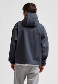 Mörkgrå huva jacka med lös passform, mjuk textur och sömdetaljer på axlarna. Bärs över en vit skjorta och grå byxor.