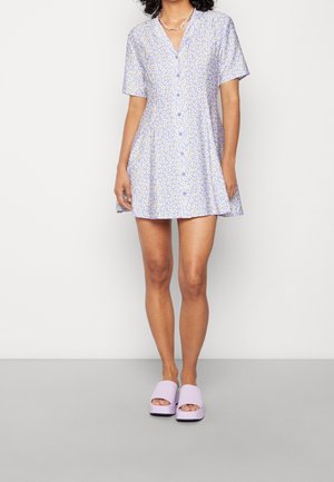 Robe lilas claire à manches courtes, motif floral jaune et blanc, encolure en V, coupe évasée. Associée à des sandales compensées lavande.