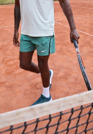 Tennis player standing on clay court wearing green shorts, white shirt, white socks, and blue sneakers, holding racket near net.