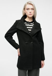 Mlada ženska v črni dvoredni plašču, oblečena v črno-bele črtaste pulover, pozira z eno roko na boku.