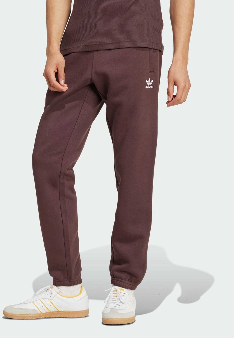 Pantalons de survêtement bordeaux avec des poignets élastiques, présentant un logo Adidas blanc sur la jambe gauche. Associés à des chaussures blanches avec des accents jaunes.