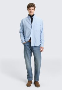 Ανοιχτό μπλε πουκάμισο με κουμπιά και τσέπη στο στήθος, φορεμένο πάνω από μαύρο ζιβάγκο, σετ με ανοιχτό μπλε τζιν και καφέ σουέντ μπότες.