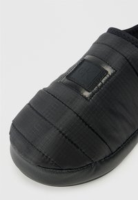 Zapatilla acolchada negra con parte superior de nylon texturizado y punta redonda. Presenta un parche con el logo en relieve y una suela de goma para mayor agarre.