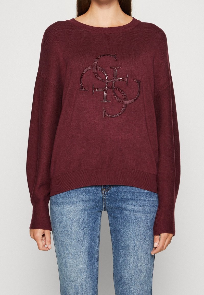 Femme portant un pull bordeaux à manches longues avec des lettres superposées ornées à l'avant, associé à un jean en denim bleu.