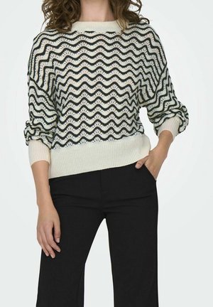 Femme portant un pantalon noir et un pull en maille blanc avec des rayures noires ondulées, une main dans la poche, debout devant un fond uni.